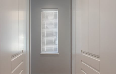 A narrow hallway with white cabinets on both sides and a window with blinds at the far end.