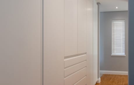 A modern, minimalist bedroom with white cabinetry and hardwood flooring, featuring a window with blinds.