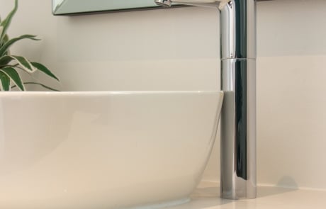 A modern chrome faucet on a minimalist bathroom sink with a white bowl, next to a mirror and a plant.