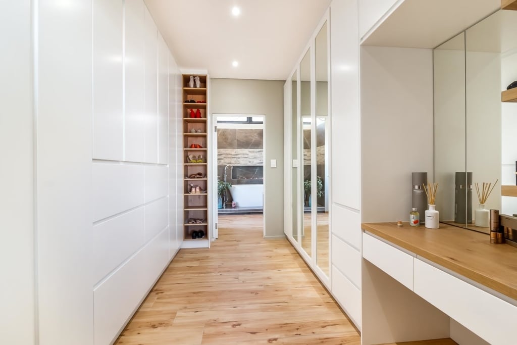 Maharaj Walk-in BIC A modern hallway with white cabinetry, a mirrored wall, and wooden flooring leading to a well-lit room.