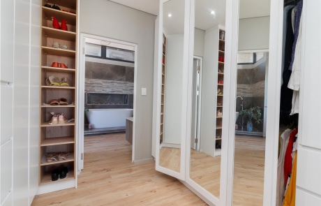 A modern closet with shelves containing shoes, a set of full-length mirrors, and a wooden floor.